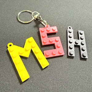 May include: A keyring featuring the letters "MSH" constructed from colourful building blocks. The letter "M" is yellow, "S" is pink, and "H" is black. The keyring includes a silver ring and clasp.