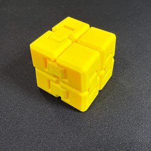 May include: A vibrant yellow fidget cube, constructed from interconnected blocks. The cube features a series of hinges, enabling it to be manipulated and folded in multiple configurations. The toy is made of a solid, smooth material.