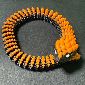 May include: A 3D-printed snake toy in a coiled position. The snake has a black body with orange segments and a detailed head. The eyes are white. The toy is designed to resemble a snake.