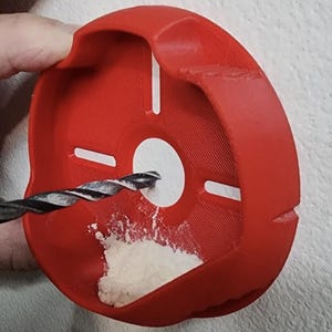 May include: A red, circular drill dust catcher with a white centre and a drill bit inserted. The catcher has a curved design with a lip and several white rectangular markings. White powder is visible inside the catcher.