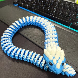 May include: A blue and white articulated snake toy. The snake's body is made of blue and white segments, with a detailed head and black eyes. The toy is designed to be flexible and poseable.