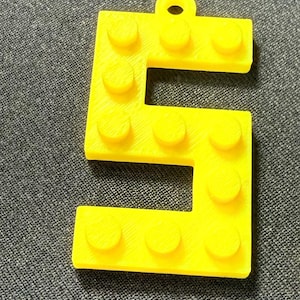May include: A vibrant yellow number 5, designed like a building block, with a small hole for hanging. It features rounded studs on its surface, characteristic of interlocking toy bricks. The background is a textured grey fabric.