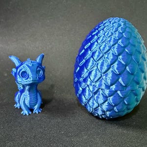 May include: A blue 3D-printed dragon figurine and a matching egg-shaped container. The dragon has large eyes and a friendly expression. The egg has a textured, scale-like pattern.
