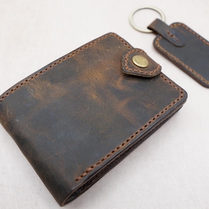 Puede incluir: Una cartera de cuero marrón con un llavero a juego. La cartera tiene un cierre de botón y está cosida con hilo marrón. El llavero es rectangular con un anillo de metal. El cuero tiene un aspecto envejecido.