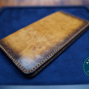Puede incluir: Cartera rectangular de cuero con acabado desgastado de dos tonos. La cartera es principalmente marrón claro con bordes marrones oscuros y está cosida con hilo en contraste. El logotipo de Aminah Leathers es visible en la esquina inferior derecha.