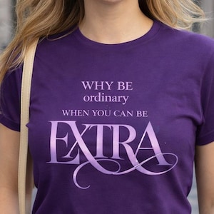 Puede incluir: Camiseta morada con el texto "WHY BE ordinary WHEN YOU CAN BE EXTRA" en una fuente estilizada. La persona lleva gafas de sol en la cabeza. Es visible una pegatina de "Printed in Europe".