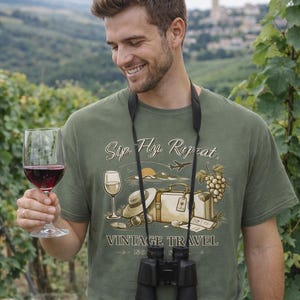 Puede incluir: Una camiseta verde oliva con el texto "Sip. Fly. Repeat." y un gráfico de viaje vintage. La imagen muestra a una persona sosteniendo una copa de vino tinto, con binoculares alrededor del cuello. Una botella y una copa de vino están sobre una barrica de madera.