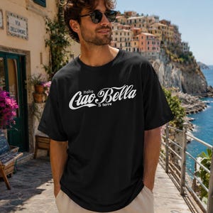 Puede incluir: Una camiseta negra de manga corta con texto blanco en el pecho que dice "Italia Ciao Bella 5 terre" en una fuente cursiva estilizada. La camiseta tiene un cuello redondo y un corte relajado, confeccionada en tela negra lisa.