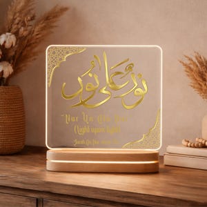 May include: A square, illuminated sign with gold Arabic calligraphy and the English text "Nur Un Ala Nur" on a light brown background. The sign sits on a wooden base and is surrounded by decorative elements.
