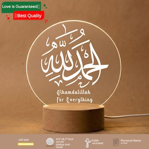 May include: A round acrylic plaque with white Arabic calligraphy and the English text "Alhamdulillah For Everything" illuminated by an LED light. The plaque is set on a wooden base, with a cable attached.