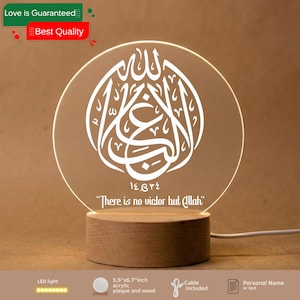 May include: A round acrylic plaque with white Arabic calligraphy, reading "There is no victor but Allah," illuminated by an LED light. The plaque is 14x17 cm and sits on a wooden base. A cable is included.
