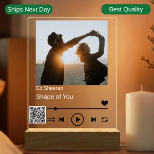 May include: An illuminated acrylic panel with a photo of a dancing couple, the song title "Shape of You" by Ed Sheeran, a QR code, and music control icons. The base is made of light-coloured wood. The panel is rectangular in shape.