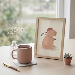 May include: A framed print of a cartoon capybara in a red hat, holding a golf club. A pink mug with a hot drink sits on a coaster, next to a pen and a small potted succulent. The scene is set on a wooden table.