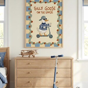 a wooden dresser with a framed poster of a cartoon goose riding a scooter on the wall above it. The poster has a playful and whimsical design, with the goose wearing sunglasses and a striped sweater. The dresser also has a small model airplane on top.