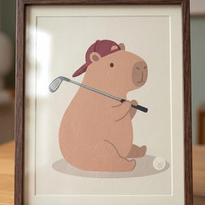 May include: A framed print showing a cartoon capybara wearing a maroon baseball cap and holding a golf club. A golf ball is on the ground. The capybara is light brown, and the frame is dark brown.