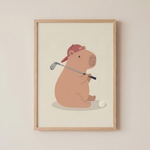 May include: A framed print of a cartoon capybara wearing a red baseball cap and holding a golf club. A white golf ball is on the ground. The capybara is light brown, and the background is a soft beige.