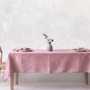 May include: A rectangular dining table covered with a solid dusty rose linen tablecloth, featuring two light wood chairs with woven seats, two white plates with folded napkins, and a small lavender ceramic vase containing a sprig of eucalyptus.