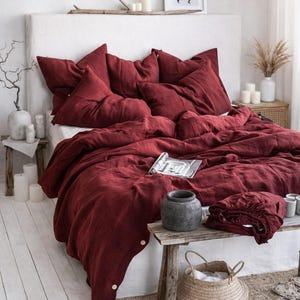 May include: A bed dressed with a deep red linen duvet cover and pillows. The bedding is on a white headboard. A small wooden bench holds a folded sheet set, a grey vase, and a woven basket. The room has a neutral colour palette.