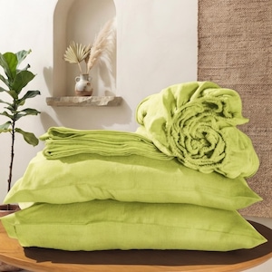 May include: A set of lime green bedding including two rectangular pillowcases stacked horizontally, a folded flat sheet, and a fitted sheet with an elasticized edge rolled into a circular bundle, all featuring a visible woven fabric texture.