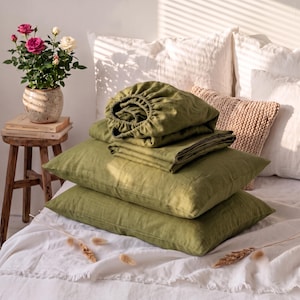 May include: A set of olive green linen bedding including two rectangular pillowcases and a folded fitted sheet. The fabric has a visible natural texture and is stacked neatly on a white bedspread. The color is a muted, earthy shade of green.