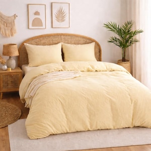 Puede incluir: Una escena de dormitorio con una cama con una colcha amarilla clara y fundas de almohada a juego. Una cabecera tejida y una mesita de noche complementan la ropa de cama. El arte mural decorativo y una planta en maceta se suman a la estética de la habitación.