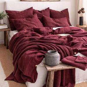May include: A bed dressed in burgundy bedding, including pillows, a duvet, and a knitted throw. A small wooden table holds a grey vase and a folded sheet set. The bed is against a white headboard and a neutral rug.