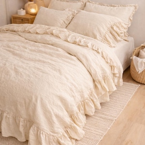 Puede incluir: Juego de ropa de cama de lino color crema con bordes con volantes. El juego incluye una funda nórdica, almohadas y una sábana bajera. La cama está sobre una alfombra tejida y se ve una mesita de noche de madera.