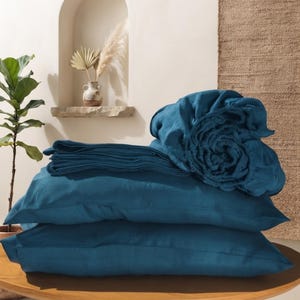 Peut inclure: Un ensemble de literie bleu sarcelle profond comprenant deux taies d'oreiller rectangulaires empilées horizontalement, un drap plat plié et un drap-housse roulé en un paquet circulaire, le tout avec une texture de tissu tissé visible.