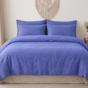 May include: A solid periwinkle blue bedding set consisting of a duvet cover and two matching rectangular pillow shams. The fabric has a visible textured weave, and the set is arranged neatly on a bed with a neutral-toned base and pillows.