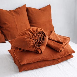 May include: A set of rust-colored bedding including two square pillow shams, a fitted sheet with elasticized edges, and a flat sheet. The fabric has a visible woven texture and a deep, earthy orange tone, presented in a stacked arrangement.