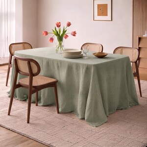 May include: A rectangular dining table covered with a sage green linen tablecloth, surrounded by four wooden chairs with woven cane backrests and seats. A clear glass vase holds pink tulips, accompanied by a stack of white plates and a glass.