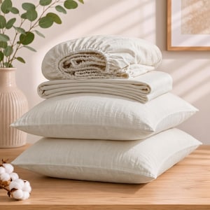 May include: A set of off-white bedding including two rectangular pillows stacked on top of each other, a folded flat sheet, and a fitted sheet with elastic edges. The fabric has a visible textured weave and a neutral, light cream color throughout.