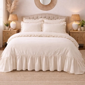 May include: A cream-colored bed set with ruffled edges, including a comforter, pillows, and a bed skirt. The bed is set against a neutral-toned wall, with matching bedside tables and lamps.