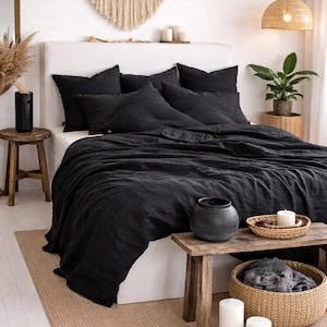 May include: A bedroom scene featuring a bed with black bedding, including a duvet cover and pillows. A woven rug and wooden furniture, including a small table and stool, complement the bed. Decorative elements include a macrame wall hanging and candles.
