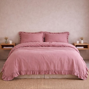 Op de afbeelding: Een stofroze beddengoedset met gerimpelde randen. De set bevat een dekbedovertrek en twee kussenslopen. Het bed is opgemaakt met een beige bedrok en staat op een geweven vloerkleed. Twee kleine houten nachtkastjes flankeren het bed.