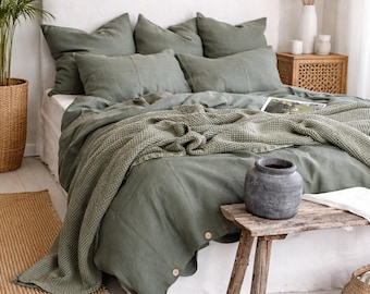Op de afbeelding: Een slaapkamer met olijfgroen linnen beddengoed, inclusief kussens en een dekbedovertrek. Een gebreide groene deken ligt op het bed. Een kleine houten tafel heeft een grijze vaas. Een geweven vloerkleed en een potplant zijn ook zichtbaar.