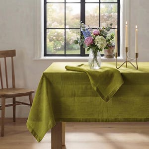 Puede incluir: Un mantel rectangular de lino verde oliva drapeado sobre una mesa, con una textura tejida visible. Un florero de vidrio estriado transparente contiene un ramo de flores rosas, blancas y azules, acompañado de un candelabro de metal dorado con tres velas.