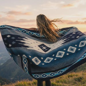 May include: A blue and white patterned blanket is held open against a mountain backdrop at sunset. The blanket features geometric designs in shades of blue, white, and grey. The blanket is being held by a person.
