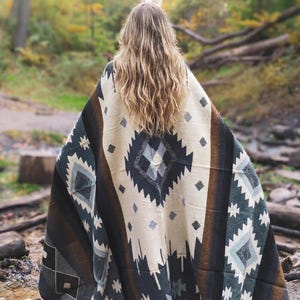 Puede incluir: Una manta estampada con un diseño geométrico en crema, negro, marrón y verde azulado. La manta está sobre alguien, de espaldas a la cámara, en un entorno forestal. El diseño de la manta presenta formas de diamantes y estrellas.