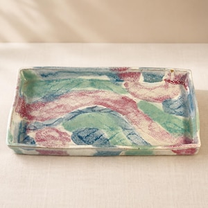 May include: A rectangular ceramic tray featuring a hand-painted abstract pattern with swirling bands of magenta, teal, and blue. The surface has a textured, speckled glaze finish with visible brushstrokes and a slightly uneven, organic edge.