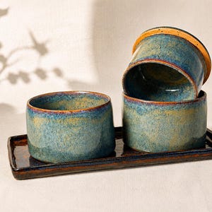 Könnte beinhalten: Drei Keramikschalen mit einer gesprenkelten blau-grünen Glasur und warmen orange-braunen Rändern, angeordnet auf einem passenden rechteckigen Keramiktablett mit dunkler, glänzender Oberfläche. Die Schalen haben eine zylindrische Form.