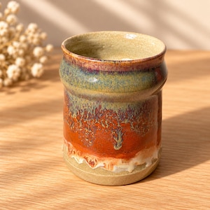 May include: A cylindrical ceramic tumbler featuring a reactive glaze with a blend of earthy tones. The upper section displays a muted green and purple hue, transitioning into a vibrant orange midsection with crystalline patterns, ending in a cream base.