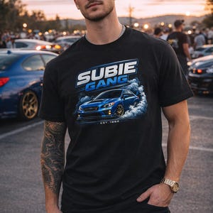 Könnte beinhalten: Schwarzes T-Shirt mit einer Grafik eines blauen Autos, das Rauch ausstößt, den Worten "SUBIE GANG" in Weiß und Blau und "EST. 1992". Das T-Shirt wird von einer Person mit einer schwarzen Kappe getragen.