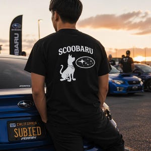 May include: Black t-shirt with the word "Scoobaru" above a white cartoon dog and a star design. The shirt is worn by a person standing near a blue Subaru car. The license plate reads "Subied".
