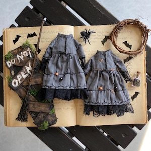 Pode incluir: Dois vestidos em miniatura com padrão xadrez preto e branco, mangas compridas, acabamento em renda preta nos punhos e na bainha, e pequenas contas laranja e pretas. Os vestidos estão sobre um livro aberto com uma placa escrita DO NOT OPEN.