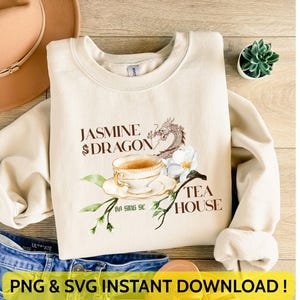 May include: A cream-coloured sweatshirt featuring the text "Jasmine Dragon Tea House" and a graphic of a teacup, dragon, and flowers. The words "PNG & SVG INSTANT DOWNLOAD!" are at the bottom. A brown hat and blue jeans are also in the image.