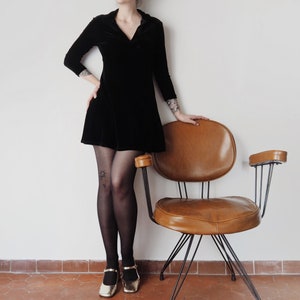 K&ouml;nnte beinhalten: Eine Frau in einem schwarzen Samtkleid und schwarzen Strumpfhosen steht neben einem braunen Ledersessel mit einem Metallgestell.