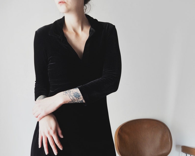 K&ouml;nnte beinhalten: Eine Frau in einem schwarzen Samtkleid mit V-Ausschnitt und langen &Auml;rmeln. Sie hat ein Blumen-Tattoo auf ihrem linken Arm.