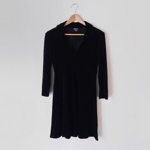 K&ouml;nnte beinhalten: Ein schwarzes Samtkleid mit Kragen und langen &Auml;rmeln. Das Kleid hat einen taillierten Oberteil und einen ausgestellten Rock.