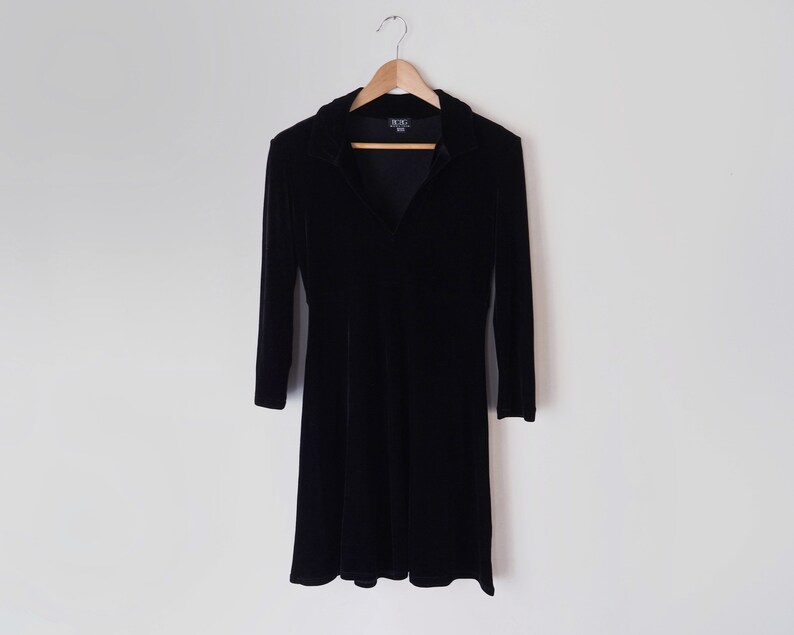 K&ouml;nnte beinhalten: Ein schwarzes Samtkleid mit Kragen und langen &Auml;rmeln. Das Kleid hat einen taillierten Oberteil und einen ausgestellten Rock.